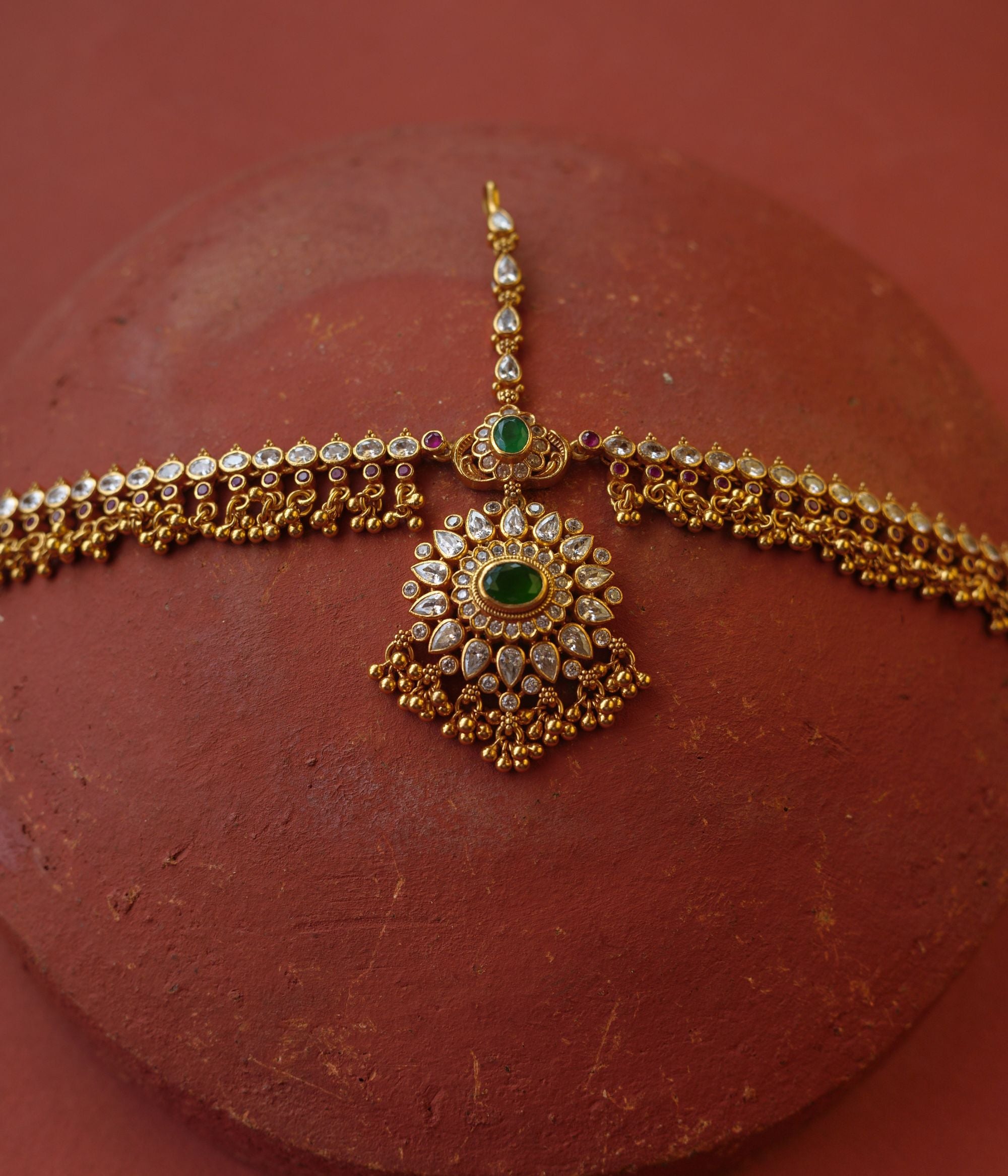 Traditional gold mathapatti with green spinal stone and Swarovski crystals in peacock eye-shaped central design.
