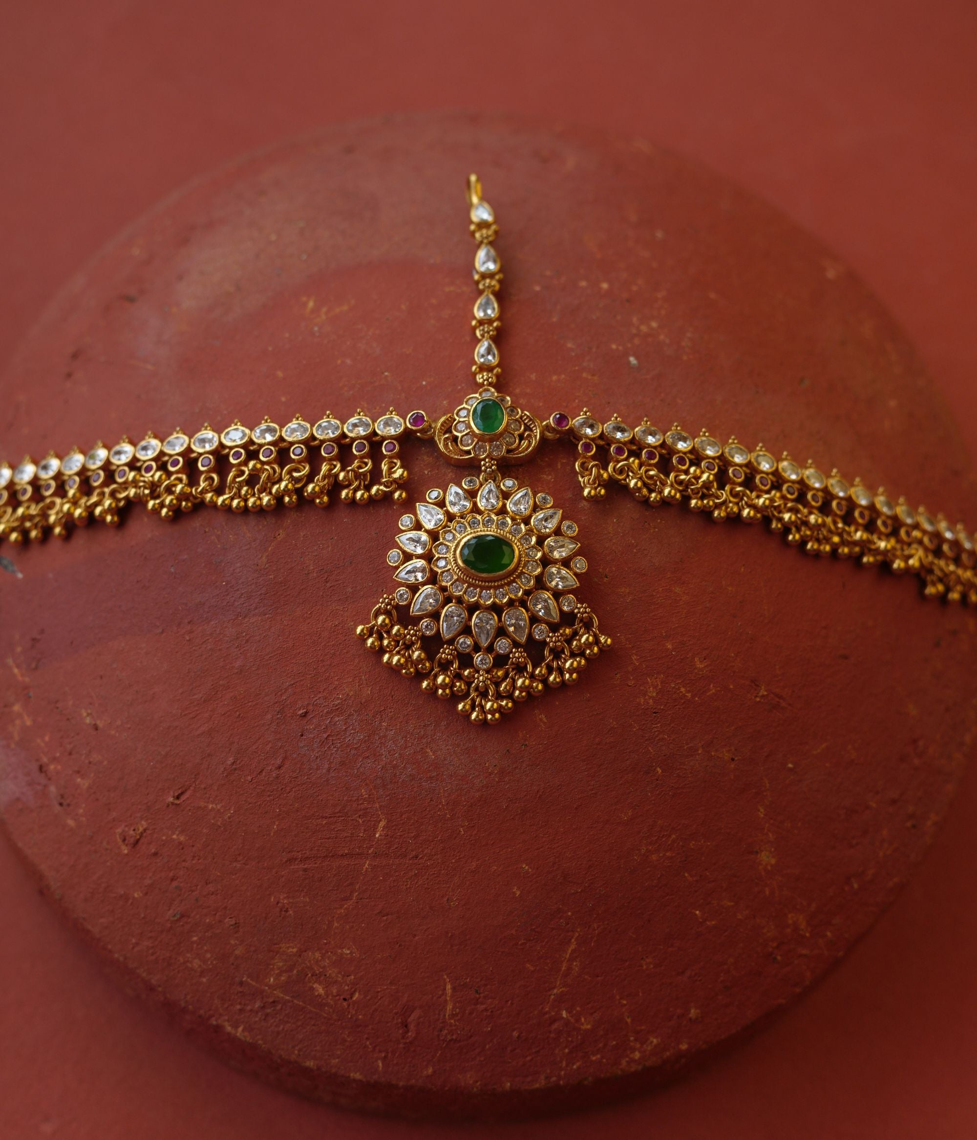 Traditional gold mathapatti with green spinal stone and Swarovski crystals in peacock eye-shaped central design.

