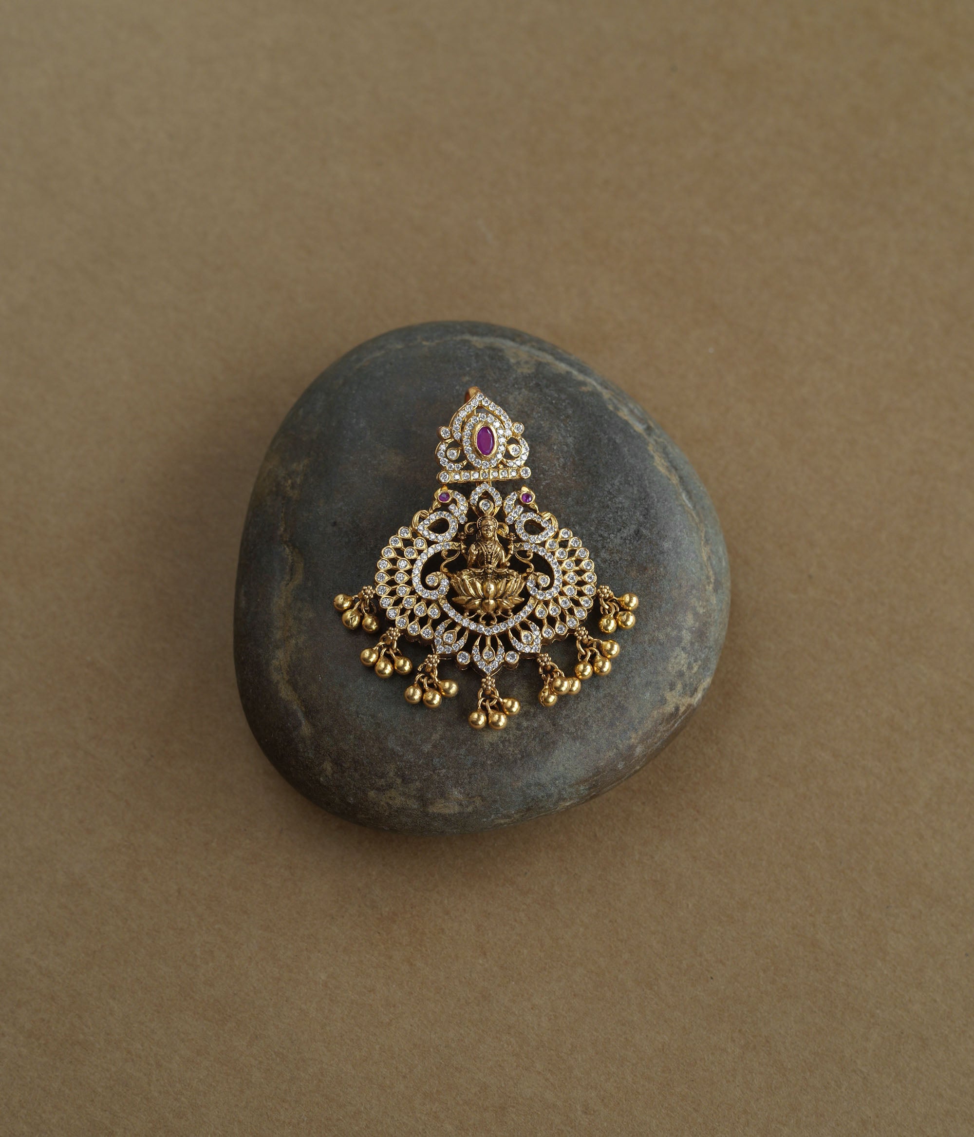 A gold pendant featuring a central deity figure. It is decorated with clear and red stones and has dangling gold beads.