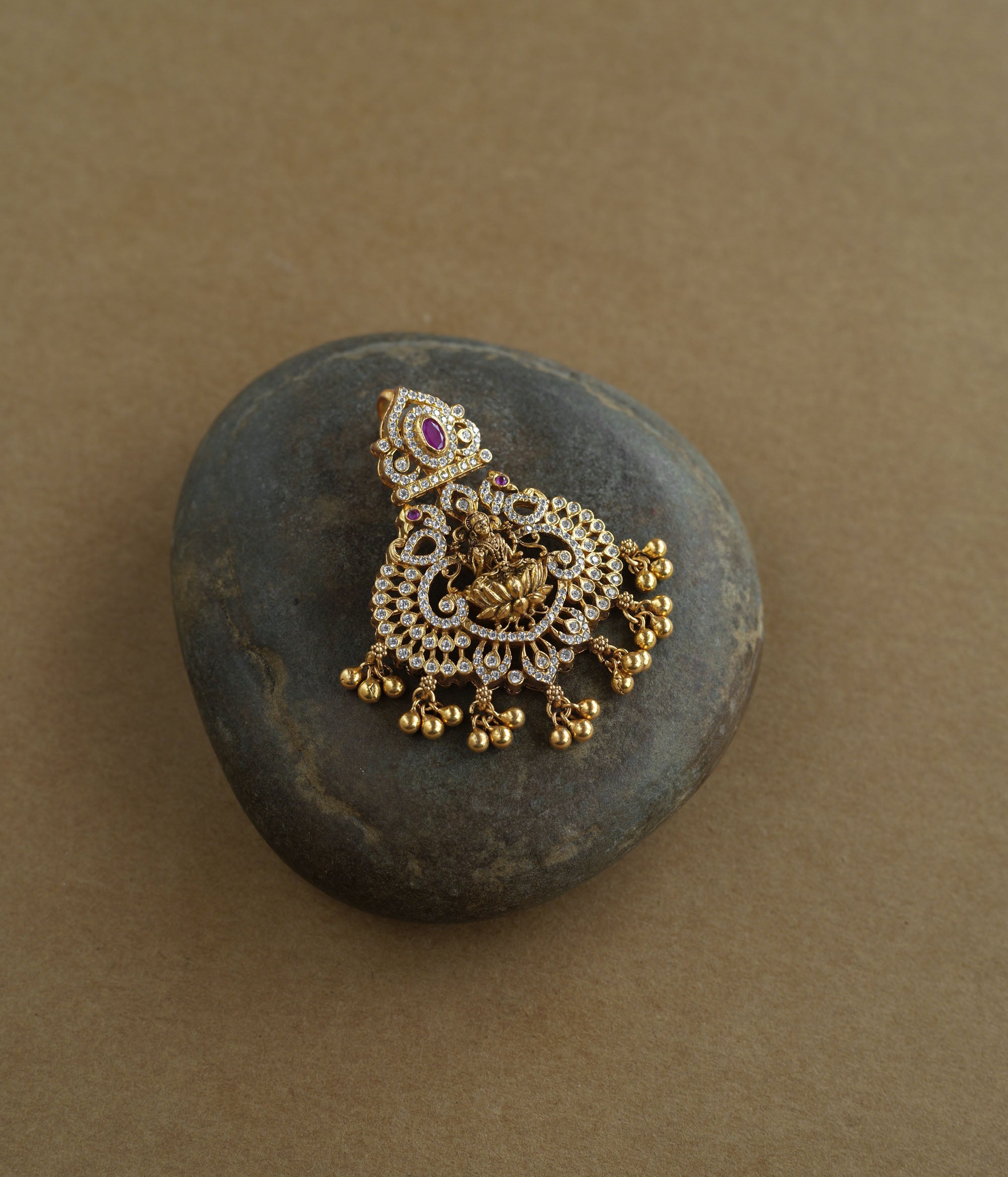 A gold pendant featuring a central deity figure. It is decorated with clear and red stones and has dangling gold beads.