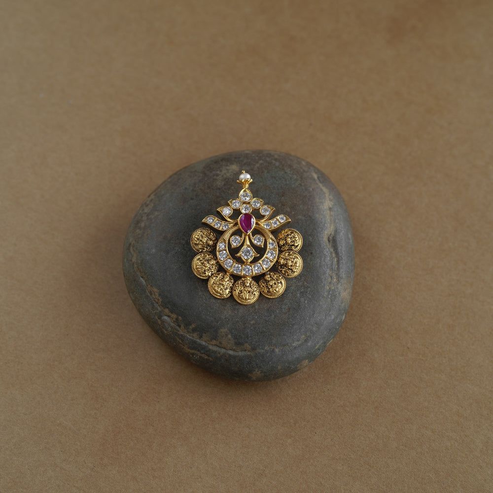 A golden pendant with a crescent-moon and floral design. It features a central red stone surrounded by clear stones and a halo of golden beads, resting on a stone.