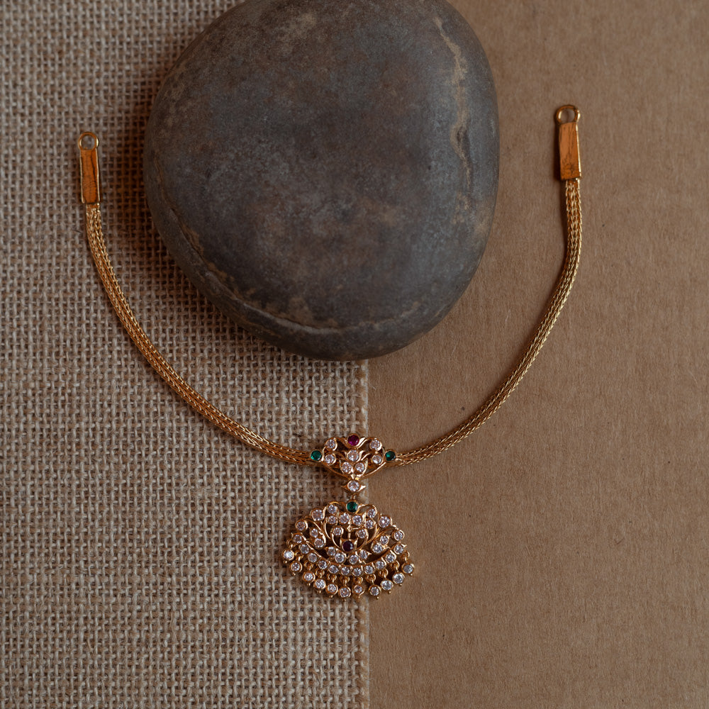 Close-up of a gold necklace with a tree-shaped pendant. The pendant is covered in clear, red, and green stones, attached to a woven gold chain.
