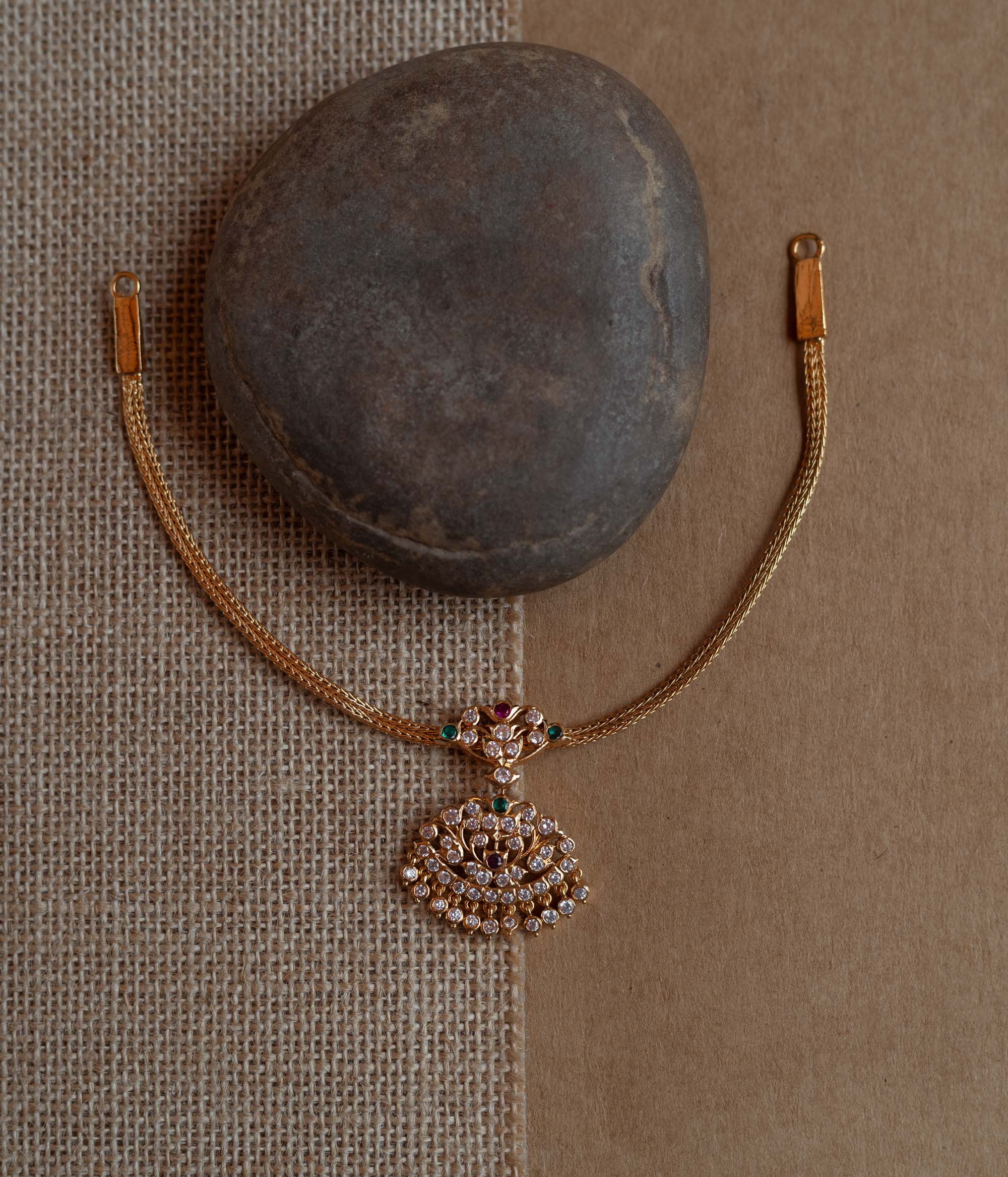 Close-up of a gold necklace with a tree-shaped pendant. The pendant is covered in clear, red, and green stones, attached to a woven gold chain.