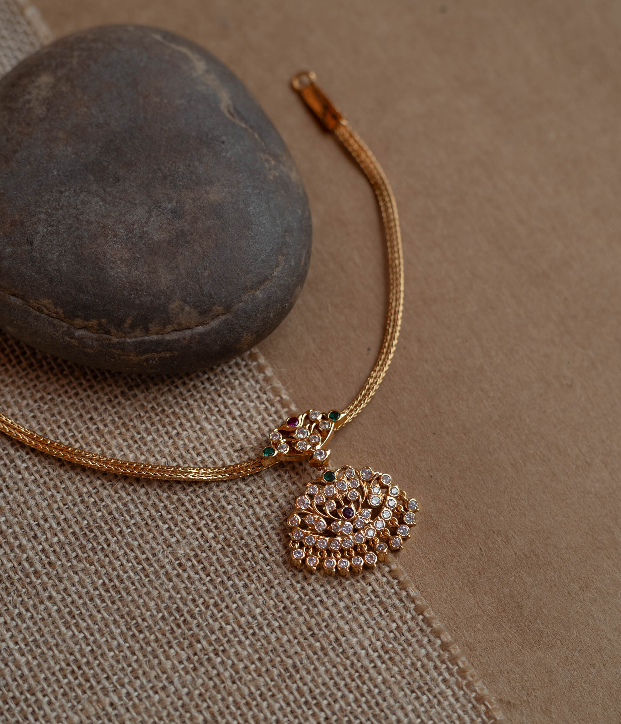 Close-up of a gold necklace with a tree-shaped pendant. The pendant is covered in clear, red, and green stones, attached to a woven gold chain.