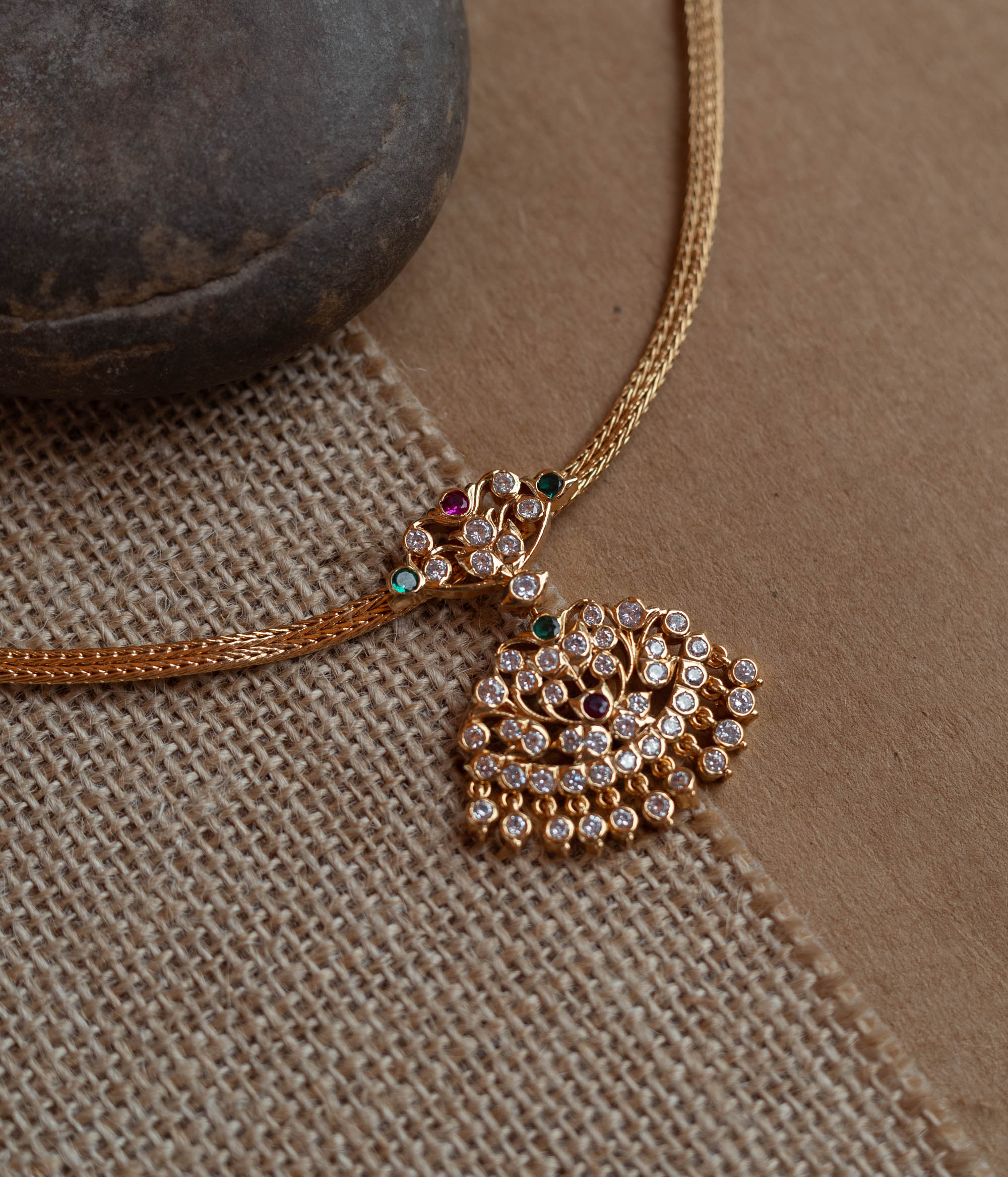 Close-up of a gold necklace with a tree-shaped pendant. The pendant is covered in clear, red, and green stones, attached to a woven gold chain.