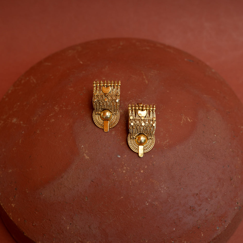 Close-up of two rectangular gold tribal-style earrings. They feature intricate textures and a prominent round gold bead at the bottom, resting on a terracotta surface.
