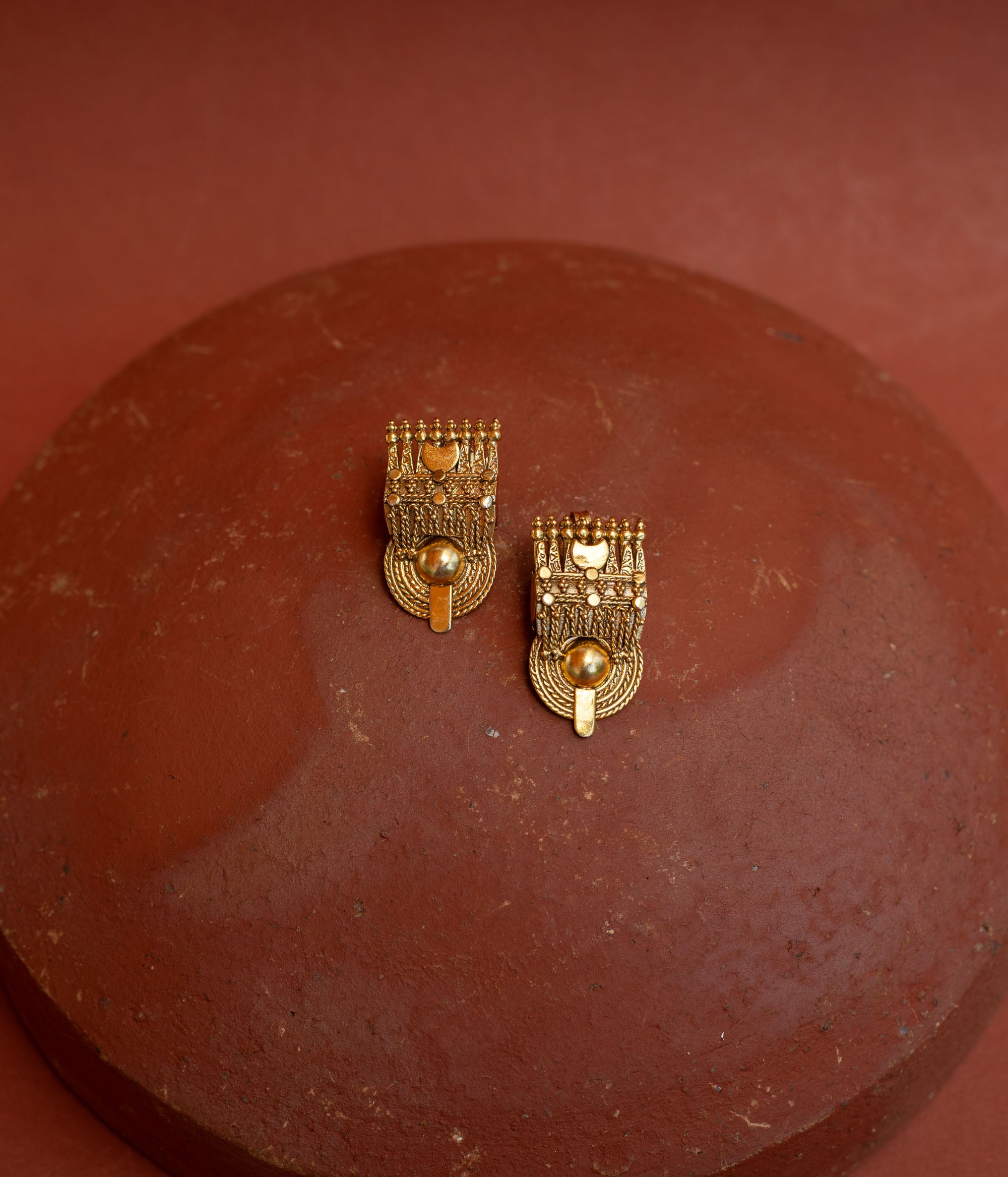 Close-up of two rectangular gold tribal-style earrings. They feature intricate textures and a prominent round gold bead at the bottom, resting on a terracotta surface.
