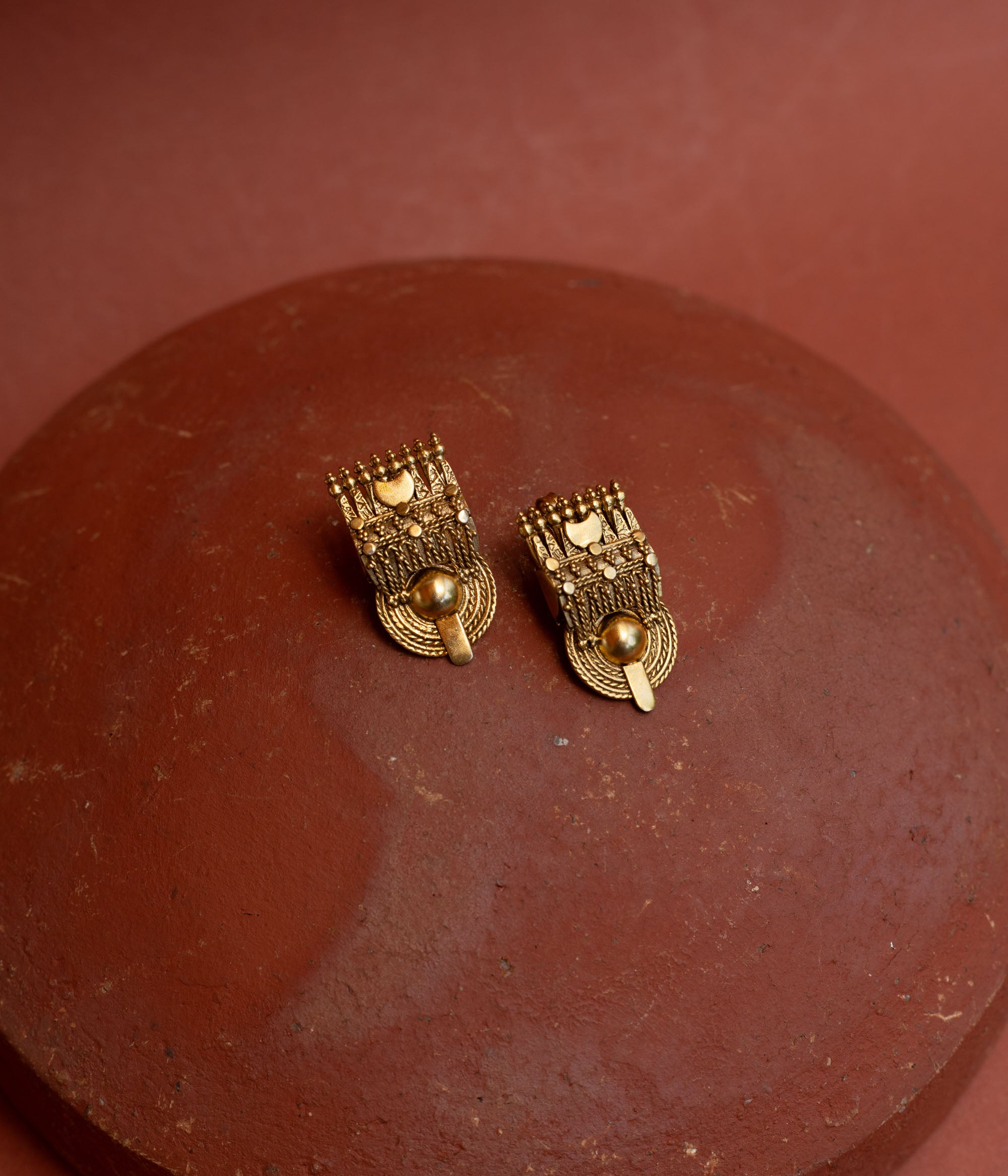 Close-up of two rectangular gold tribal-style earrings. They feature intricate textures and a prominent round gold bead at the bottom, resting on a terracotta surface.
