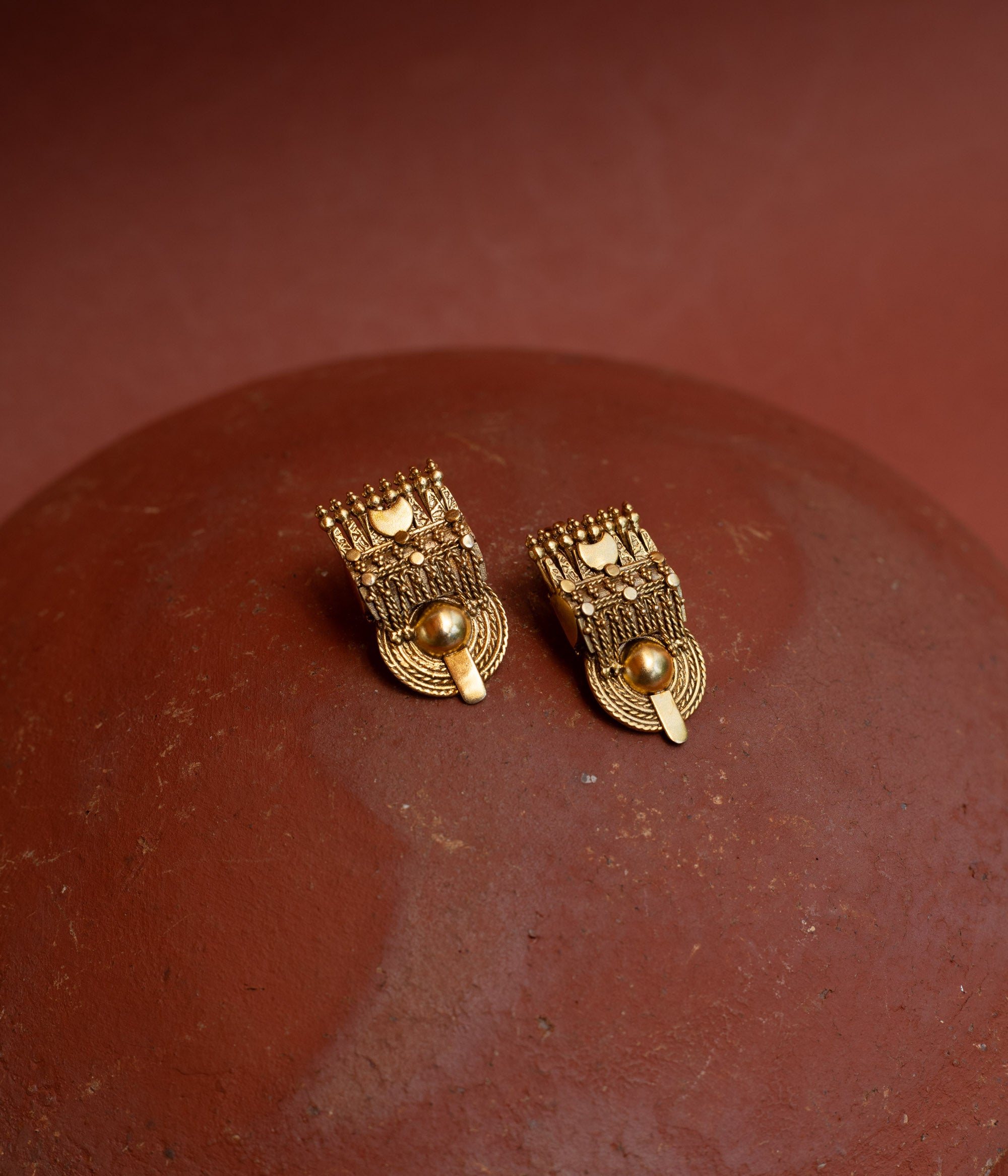 Close-up of two rectangular gold tribal-style earrings. They feature intricate textures and a prominent round gold bead at the bottom, resting on a terracotta surface.
