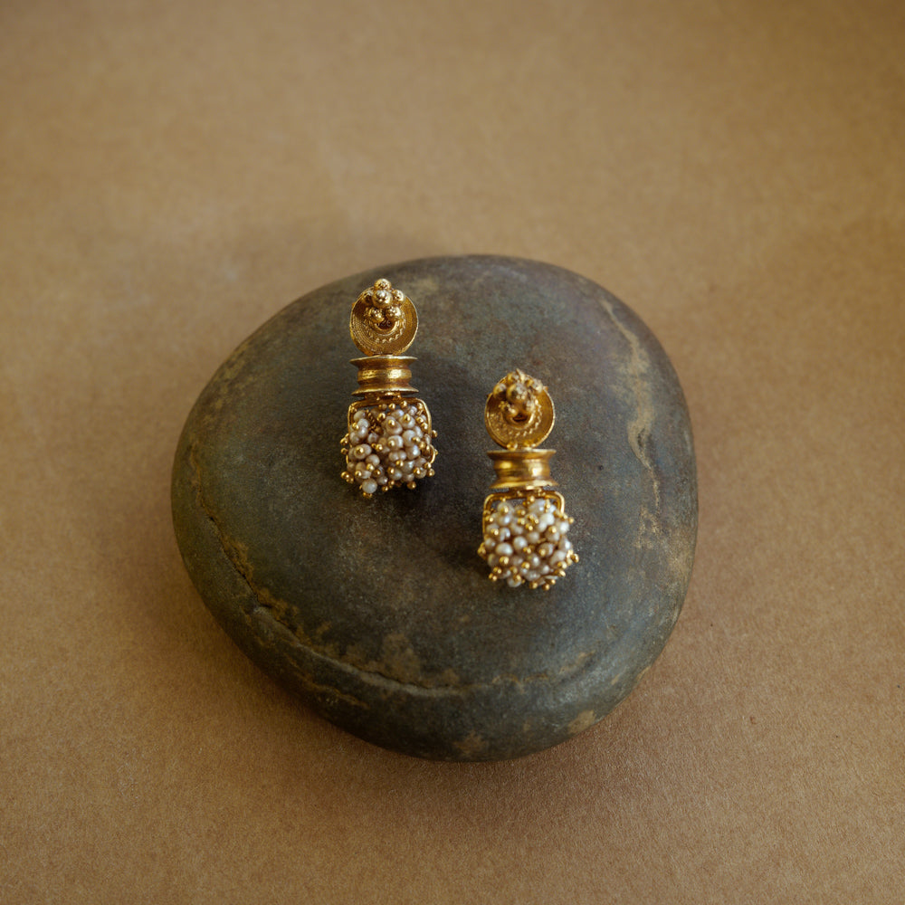 Gold earrings with a cylindrical pillar-like shape. A cluster of small white pearls hangs from the base. The earrings are on a textured brown surface.
