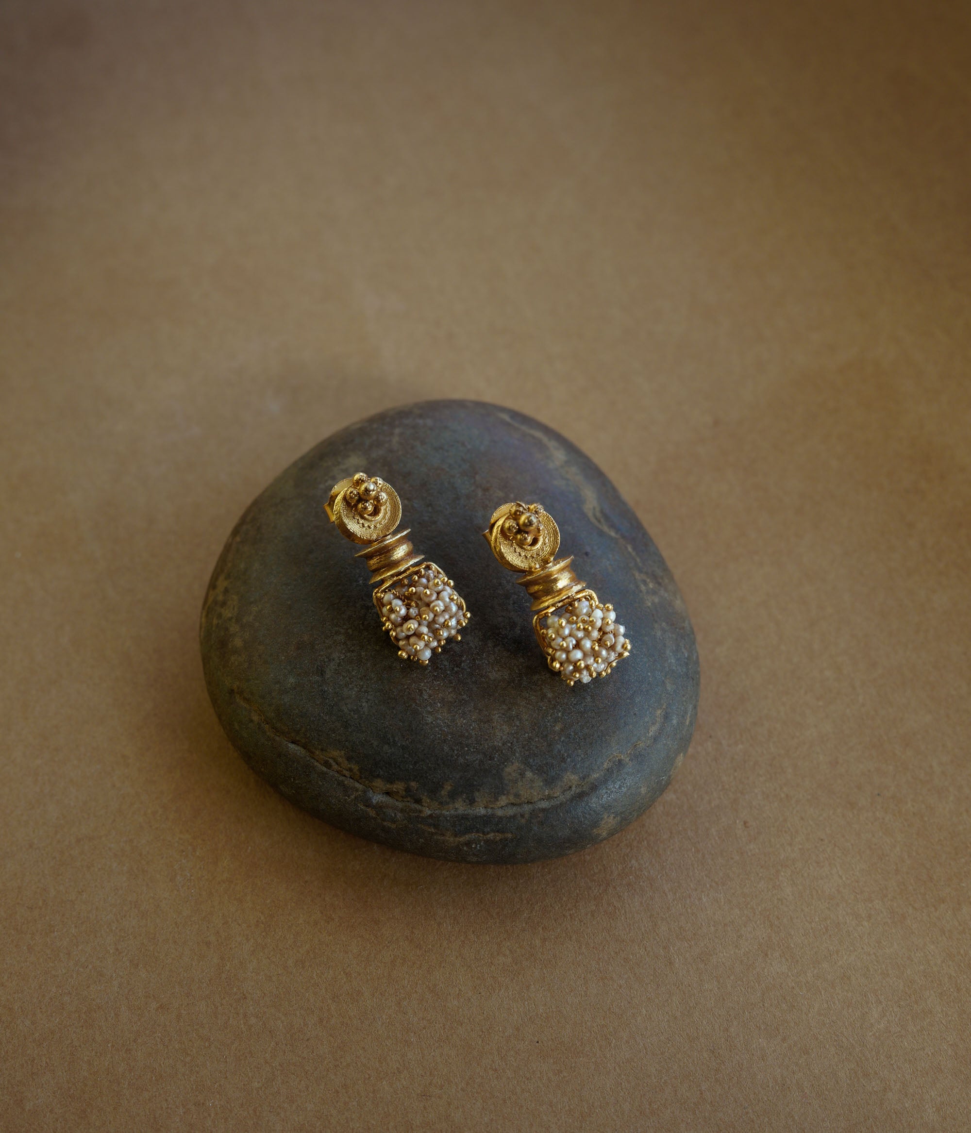Gold earrings with a cylindrical pillar-like shape. A cluster of small white pearls hangs from the base. The earrings are on a textured brown surface.