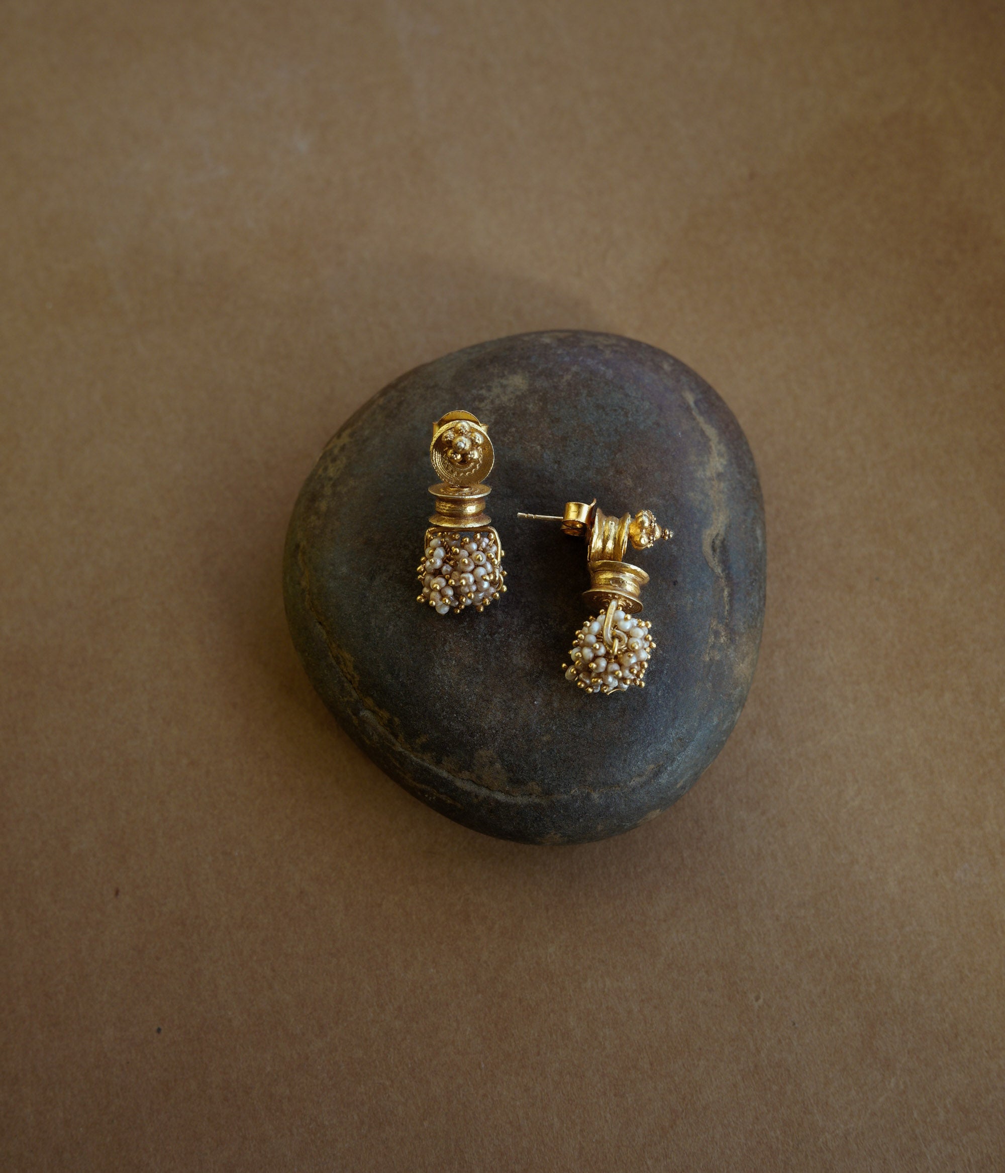 Gold earrings with a cylindrical pillar-like shape. A cluster of small white pearls hangs from the base. The earrings are on a textured brown surface.