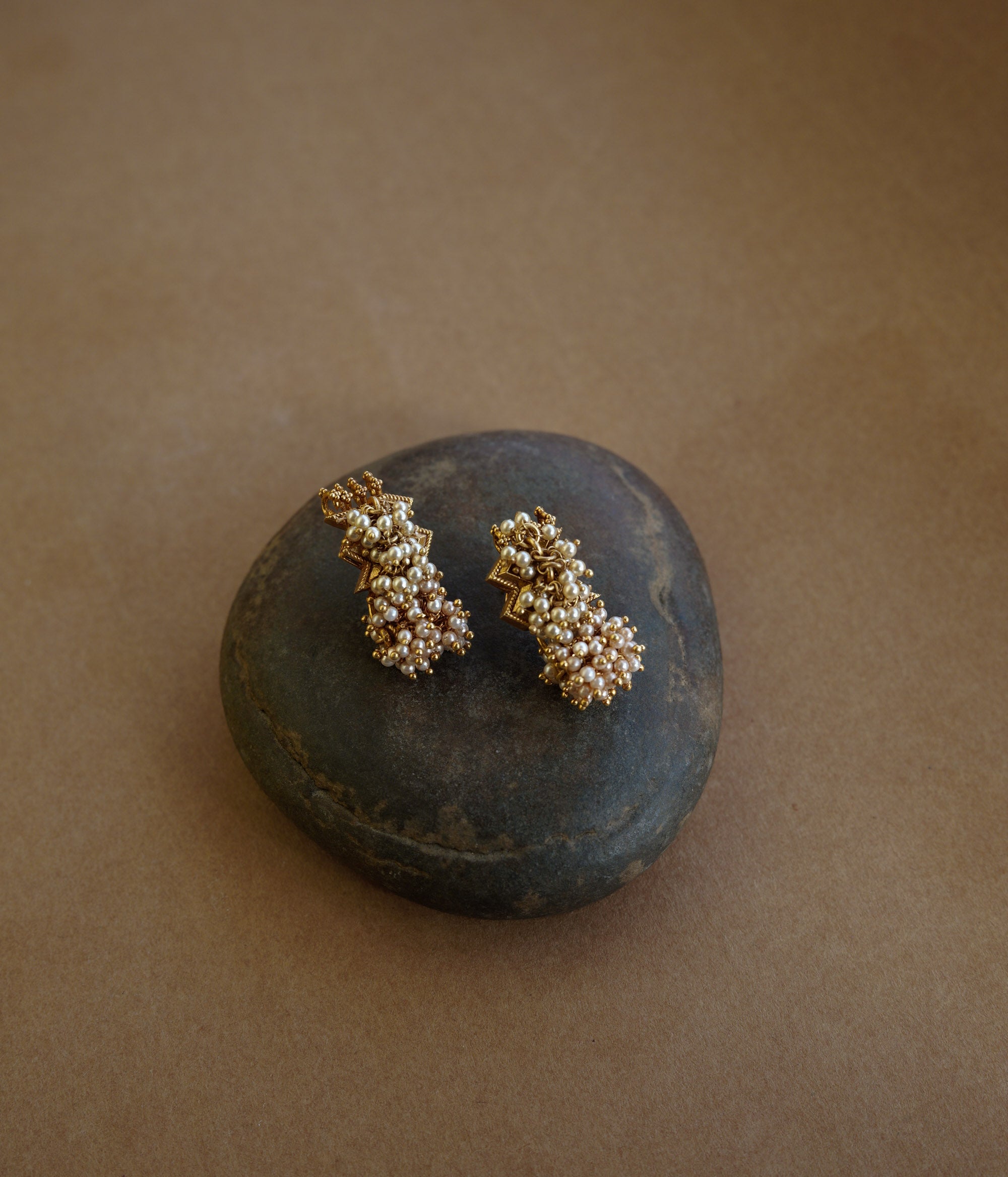 Gold earrings with a jagged, star-like motif at the top and a cluster of golden-hued beads/pearls dangling below. The earrings are on a textured brown surface.
