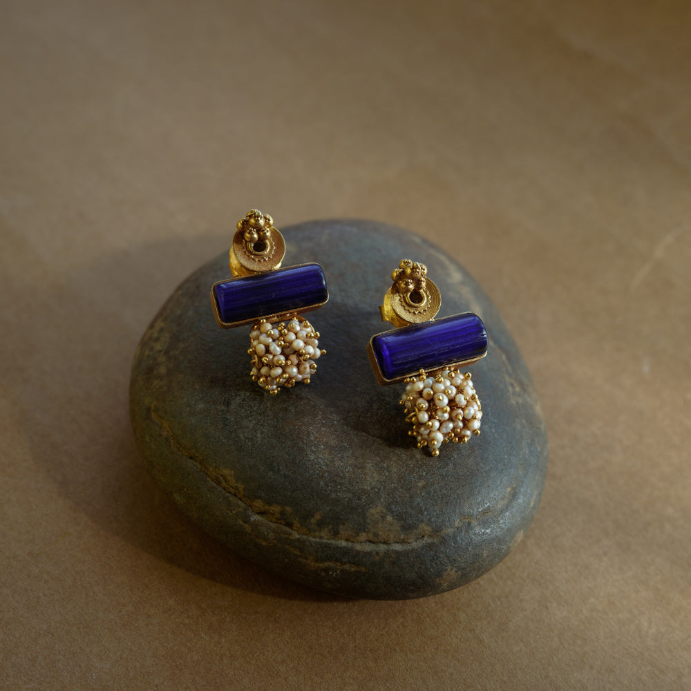 Gold earrings with a rectangular blue stone at the top and a cluster of small white pearls dangling below, resting on a textured brown surface.

