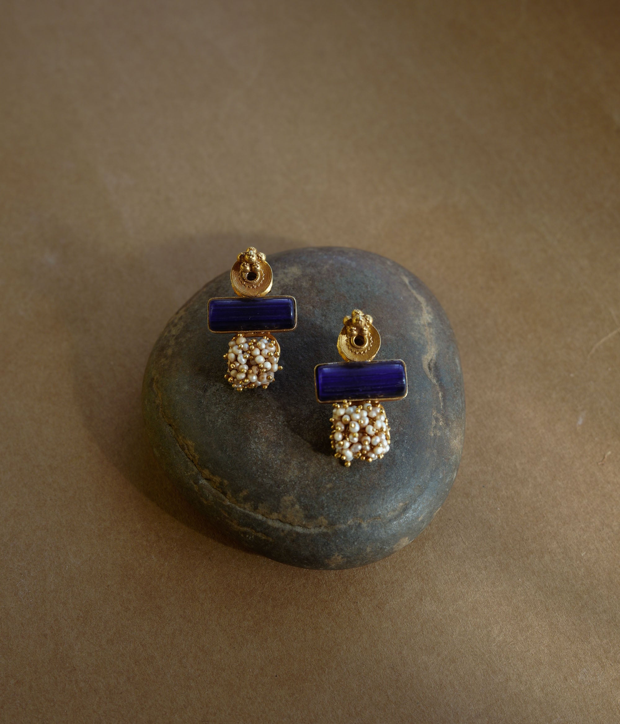 Gold earrings with a rectangular blue stone at the top and a cluster of small white pearls dangling below, resting on a textured brown surface.