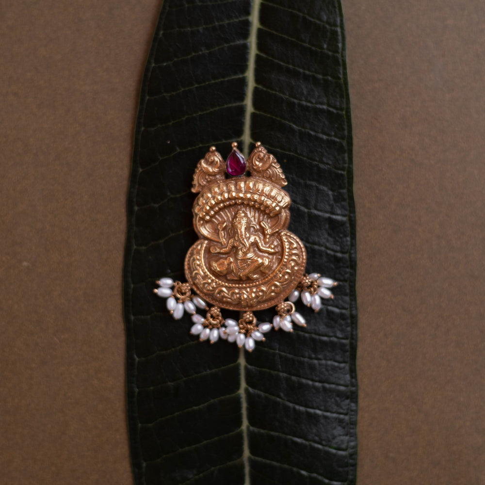 Antique gold Ganesha pendant with a conch-like top, red spinel, and pearl drops, on a dark green leaf.