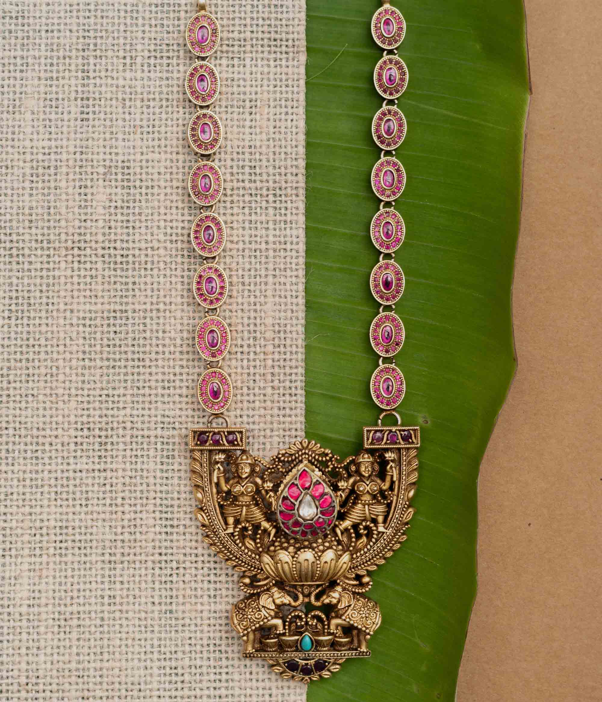 A 92.5 sterling silver GajaLakshmi Temple Heritage Necklace, likely gold-plated, featuring a pendant with Goddess Lakshmi flanked by elephants, embellished with ruby pink stones and a central kundan teardrop, with an intricate chain of oval motifs exuding vintage charm, ideal for bridal and traditional ensembles rooted in South Indian heritage.