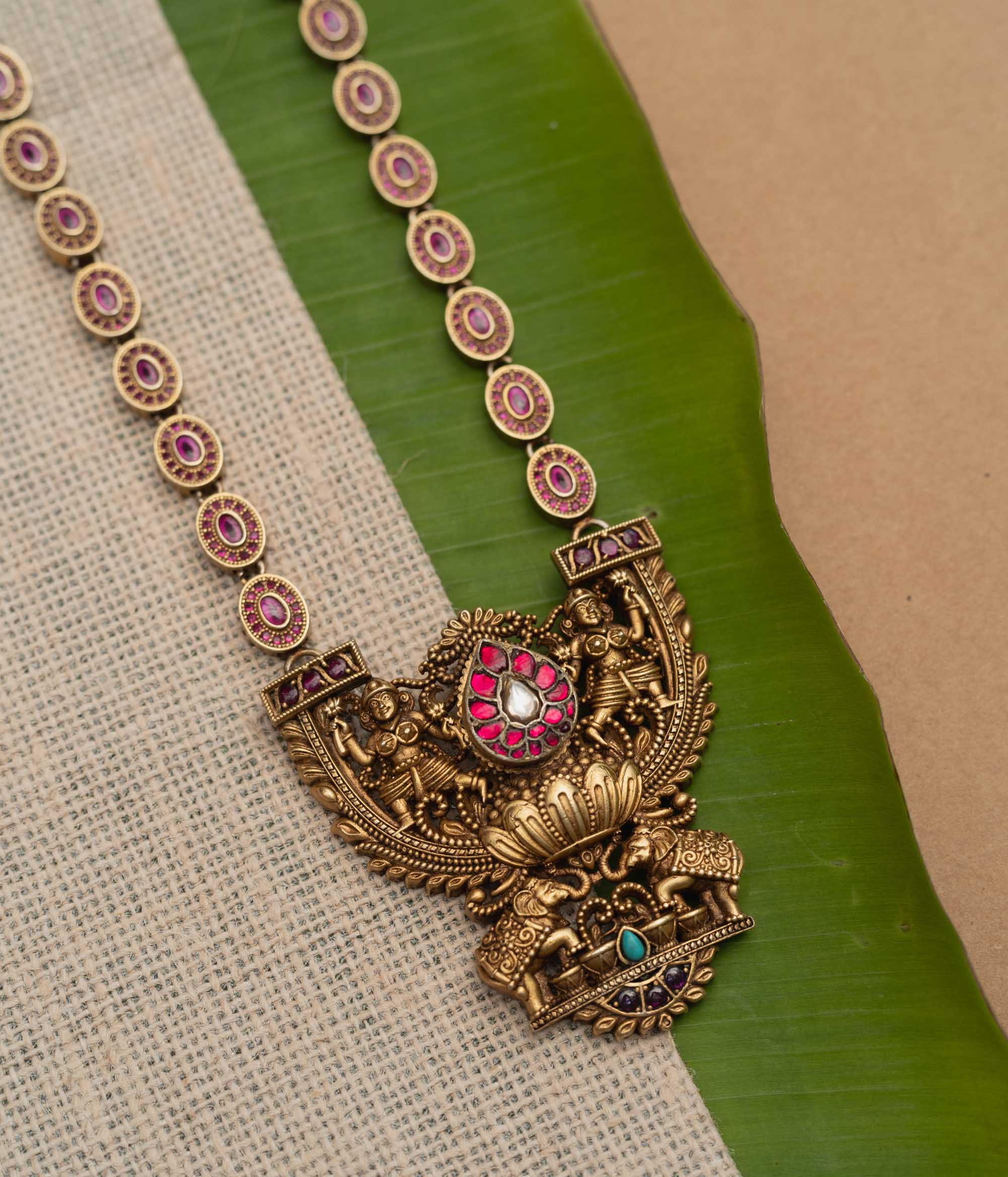 A 92.5 sterling silver GajaLakshmi Temple Heritage Necklace, likely gold-plated, featuring a pendant with Goddess Lakshmi flanked by elephants, embellished with ruby pink stones and a central kundan teardrop, with an intricate chain of oval motifs exuding vintage charm, ideal for bridal and traditional ensembles rooted in South Indian heritage.