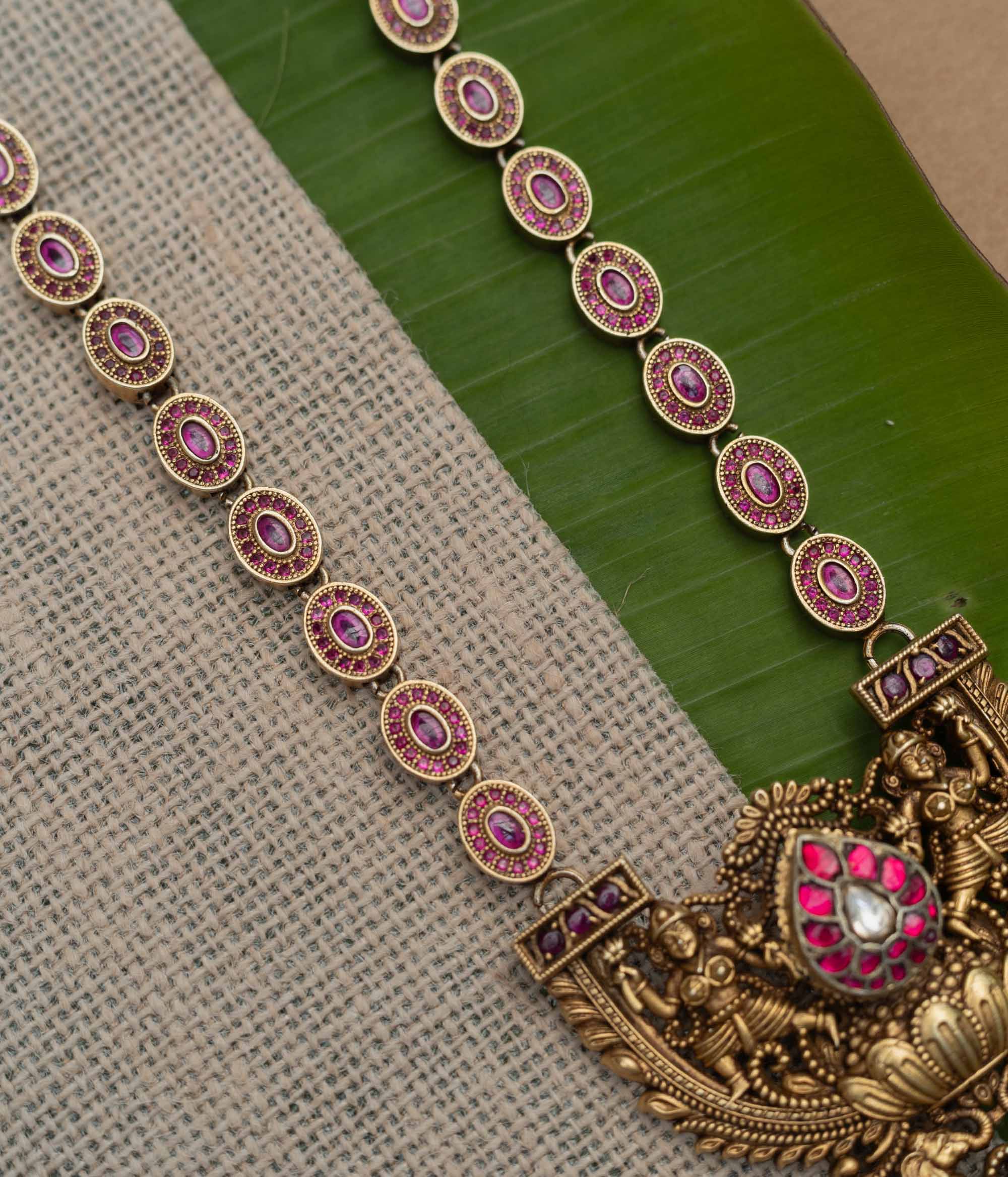 A 92.5 sterling silver GajaLakshmi Temple Heritage Necklace, likely gold-plated, featuring a pendant with Goddess Lakshmi flanked by elephants, embellished with ruby pink stones and a central kundan teardrop, with an intricate chain of oval motifs exuding vintage charm, ideal for bridal and traditional ensembles rooted in South Indian heritage.