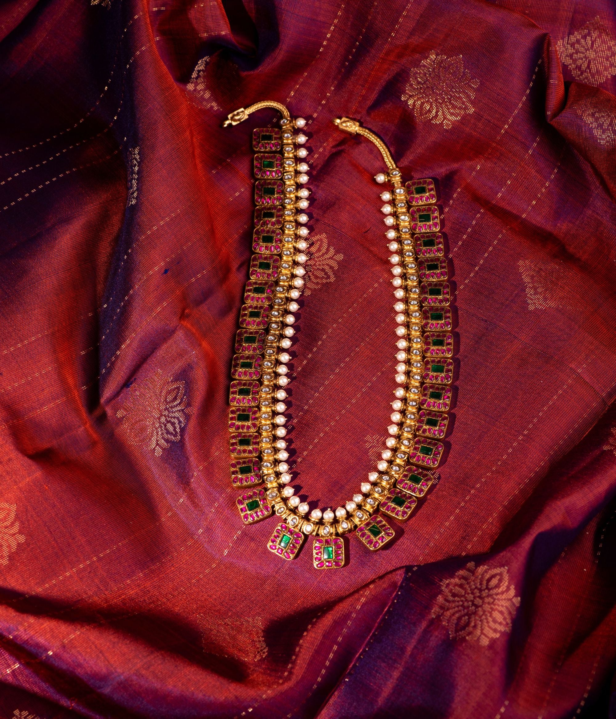 Exquisite gold-plated silver necklace featuring a unique chain composed of square-shaped drops, each adorned with vibrant red and green Kundan stones. The elegant design is further enhanced by a row of white Kundan stones and delicate pearls gracefully holding the chain, adding a regal and timeless charm to the piece.