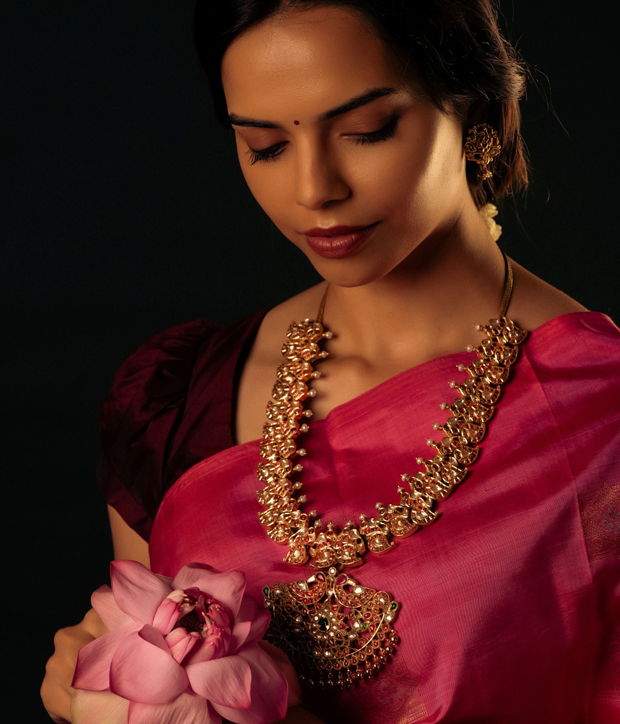 Detailed view of an antique silver, gold-plated necklace featuring a central floral-designed pendant adorned with red Kundan stones, white Kundan stones, and green Kundan stones. The necklace showcases intricate peacock motifs and mango-shaped designs embedded with stones, adding to its regal charm. Smaller pendants dangle along the lower edge, while white pearls and golden beads enhance its elegance, making it a perfect blend of tradition and sophistication.