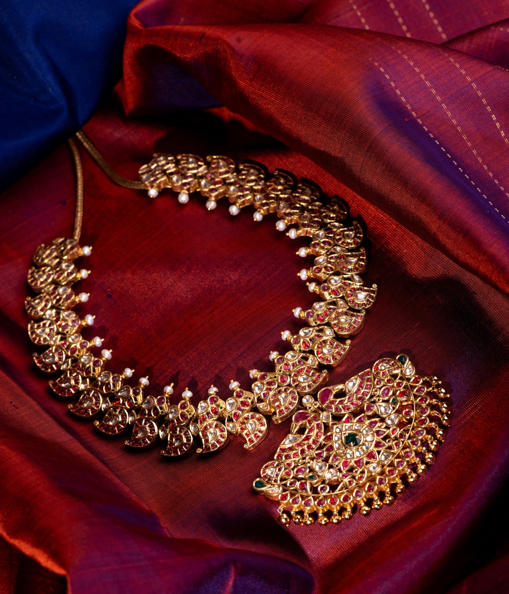 Detailed view of an antique silver, gold-plated necklace featuring a central floral-designed pendant adorned with red Kundan stones, white Kundan stones, and green Kundan stones. The necklace showcases intricate peacock motifs and mango-shaped designs embedded with stones, adding to its regal charm. Smaller pendants dangle along the lower edge, while white pearls and golden beads enhance its elegance, making it a perfect blend of tradition and sophistication.