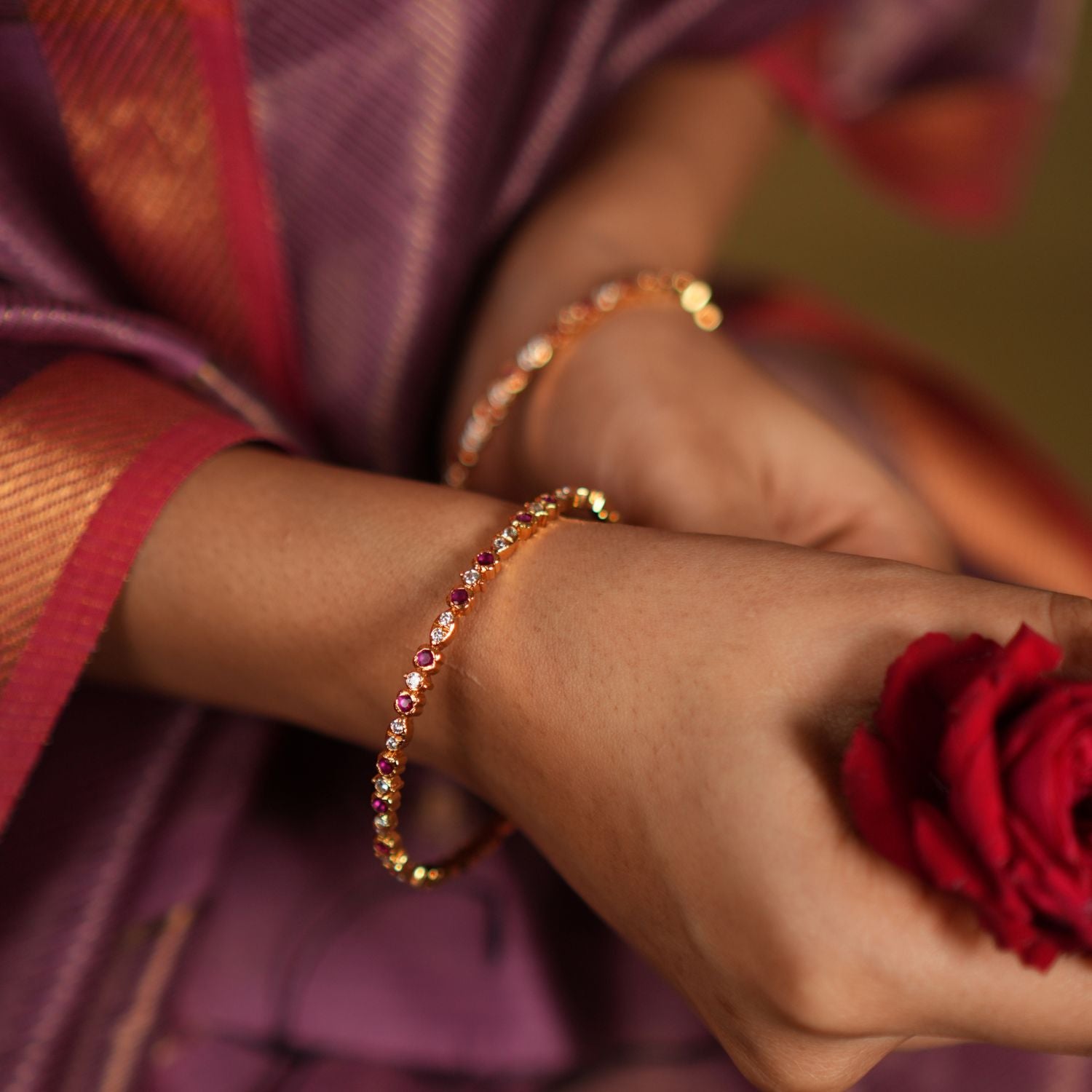 Close-up of a woman's wrist adorned with elegant silver bangles, gold-plated and embellished with white and red cubic zirconia stones, complemented by a vibrant red rose and a traditional silk saree in shades of purple and red. 