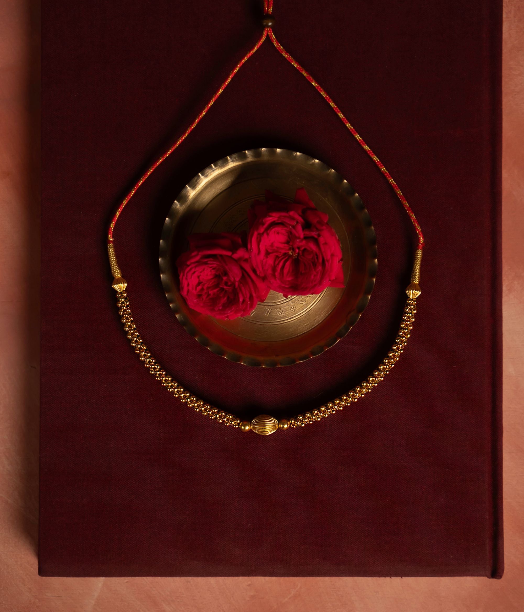 Gold-plated silver necklace featuring a delicate arrangement of small gold beads with a larger, textured gold bead at the center. Displayed on deep red fabric, accompanied by a brass bowl and red roses in the background, exuding elegance and traditional charm.
