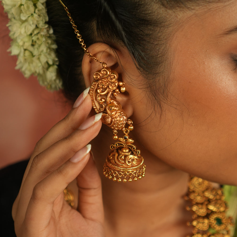 Regal peacock reborn! This gold-plated temple jhumka features intricate peacock motifs and a vibrant red spinel stone, with a delicate ear chain of tiny golden beads. A timeless blend of avian artistry and cultural opulence.