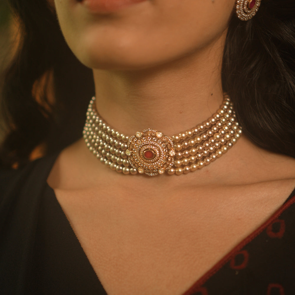 A gold choker featuring multiple strands of white pearls and a central sunburst-style medallion with red and white stones, worn on a person's neck.