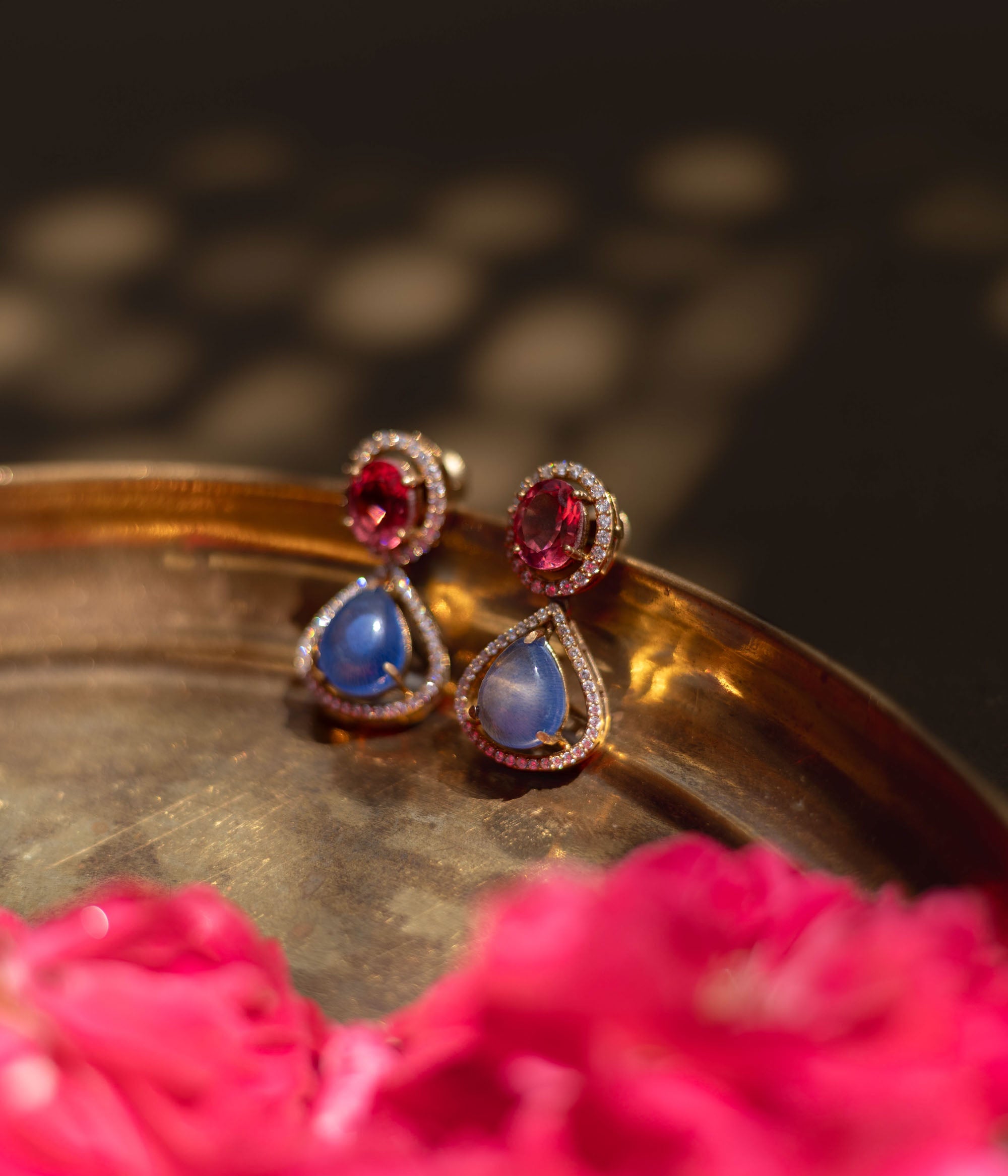 Close-up of Victorian-style teardrop earrings. Featuring pink and blue gemstones with diamond halos, resting on a textured gold surface with floral accents.