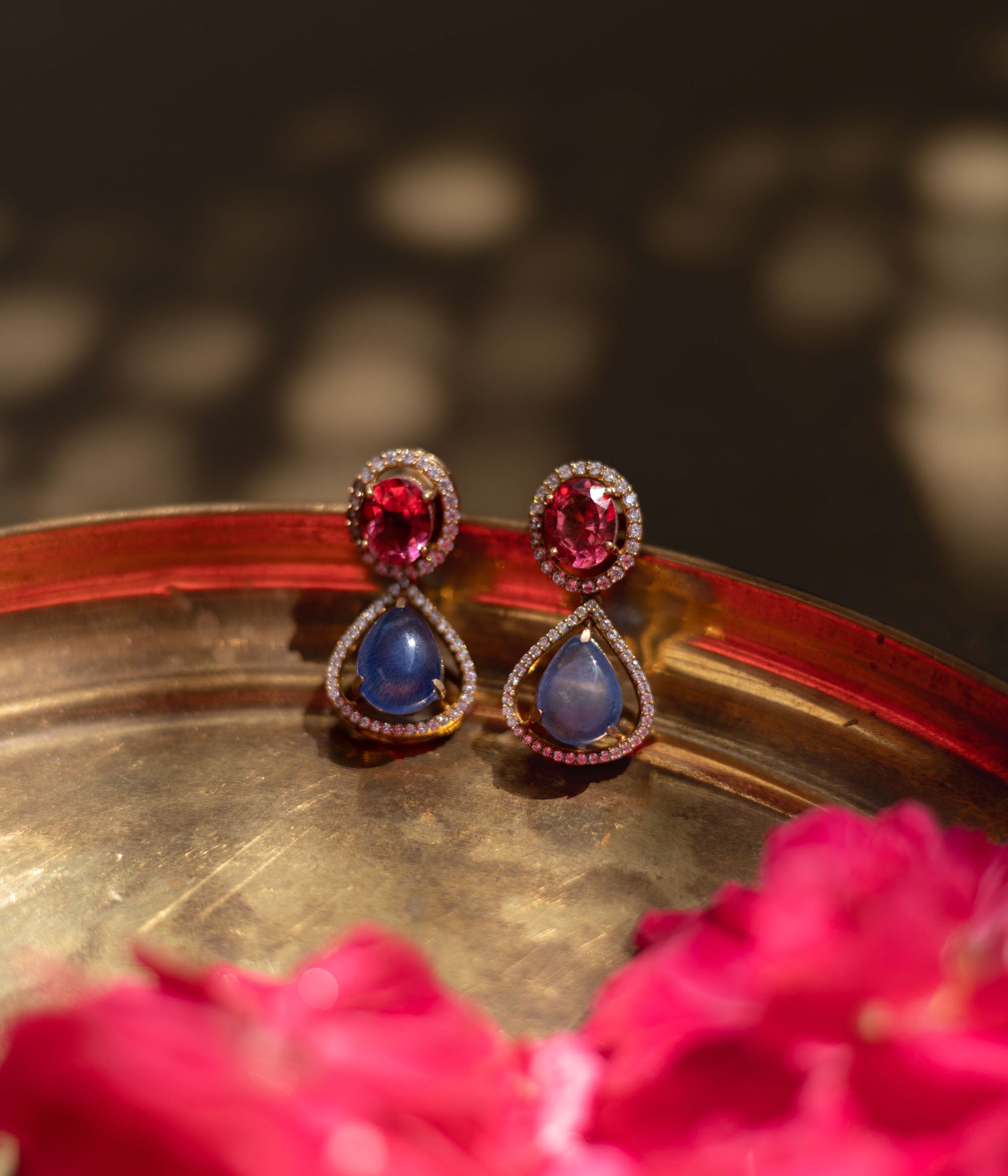 Close-up of Victorian-style teardrop earrings. Featuring pink and blue gemstones with diamond halos, resting on a textured gold surface with floral accents.