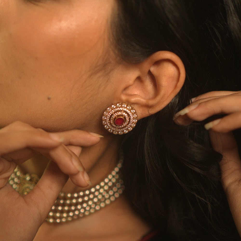 A pair of round stud earrings on a woman's ear, featuring a central red stone surrounded by concentric circles of white stones.