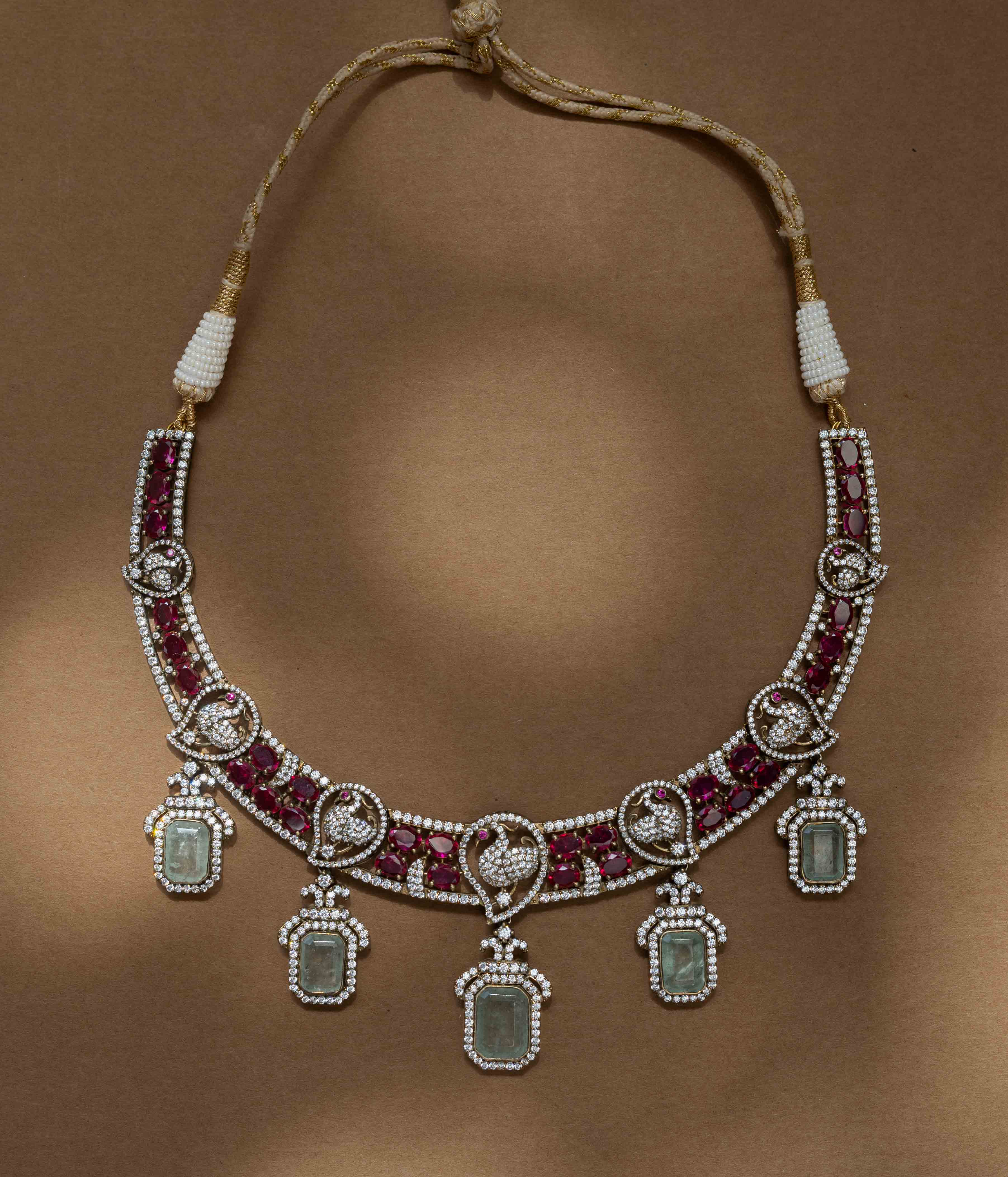 A two-tiered necklace with a large pendant featuring a green gem, clear and red stones, and pearl drops.