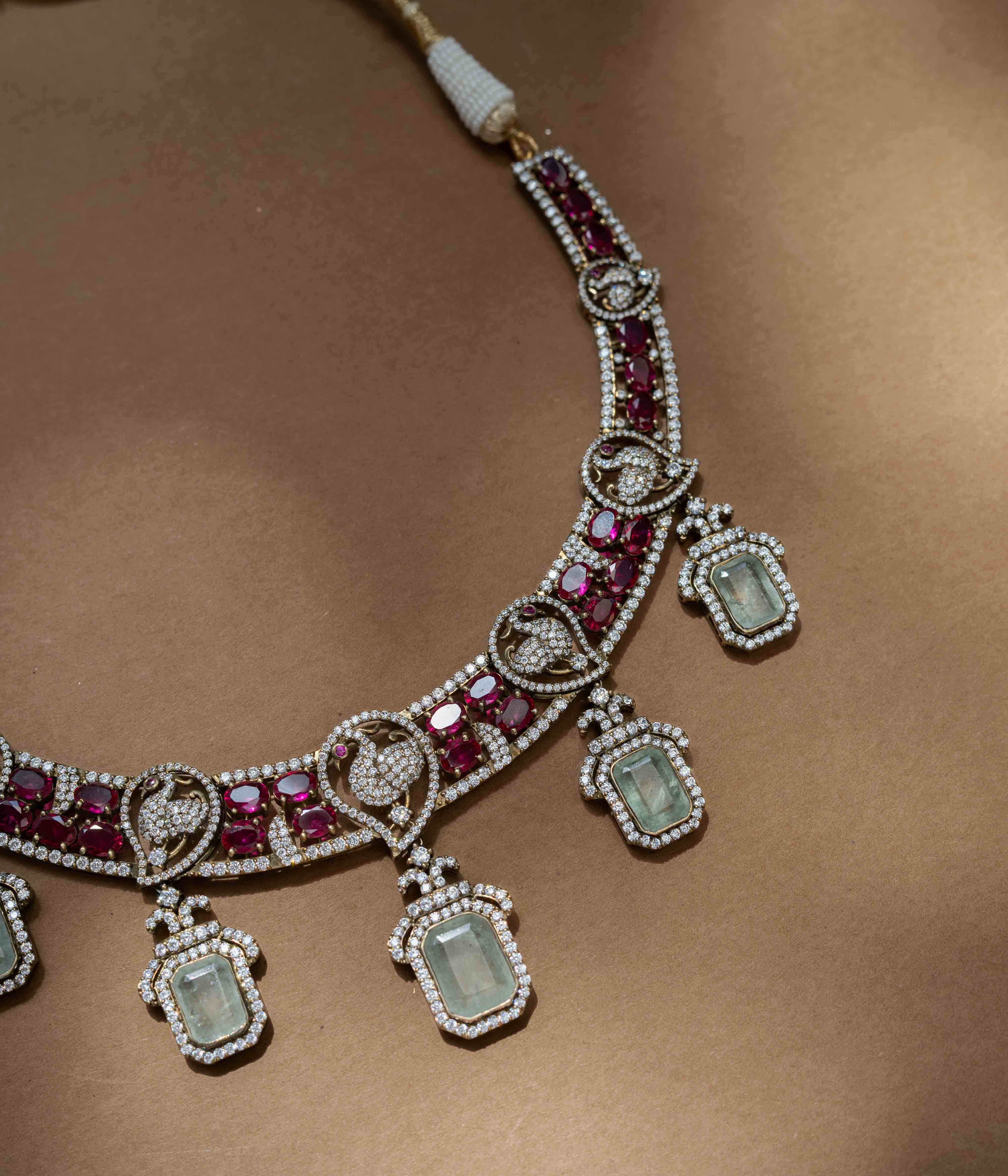 A two-tiered necklace with a large pendant featuring a green gem, clear and red stones, and pearl drops.