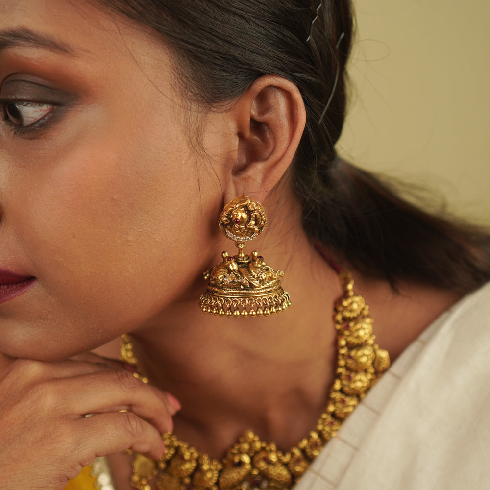 Regal grandeur meets timeless artistry! These gold-plated silver jhumkas, adorned with temple and peacock motifs, shimmer with red spinel and CZ stones, blending heritage craftsmanship with contemporary elegance.