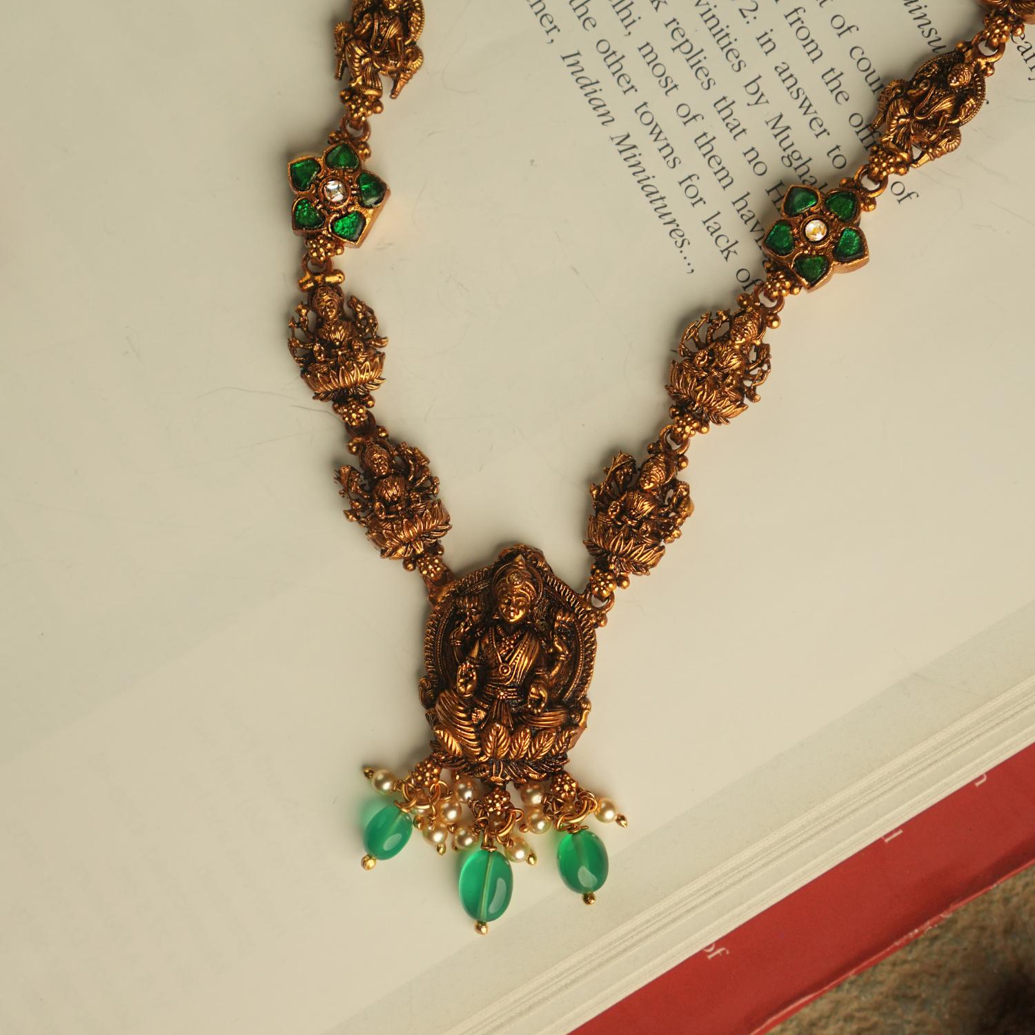 The Aadhya necklace evokes the charm of a bygone era, with antique gold, polkis, and a divine Lakshmi pendant. Pearl drops and green beads add a touch of timeless grace.