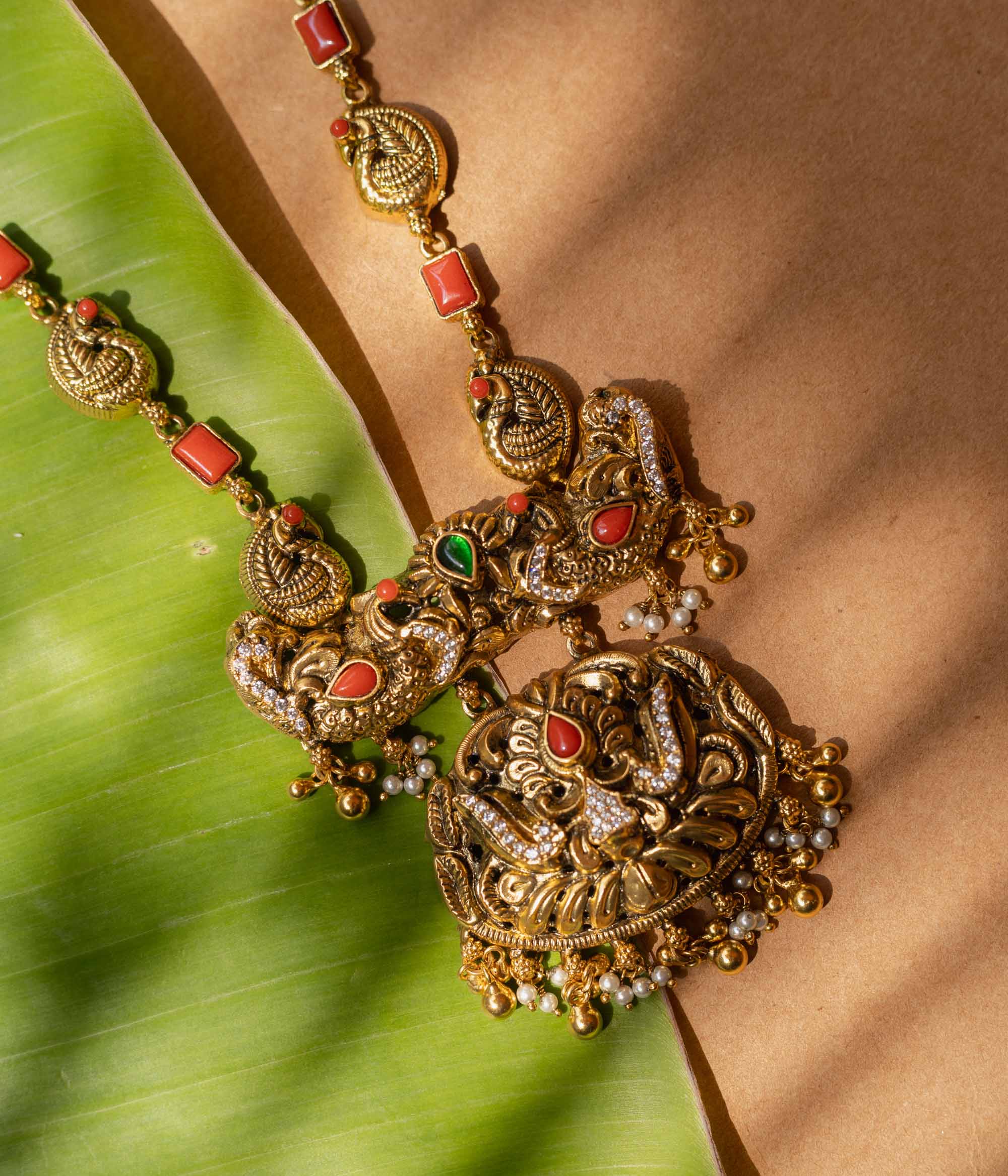 A 92.5 sterling silver Mythical Peacocks Antique Temple Necklace, likely gold-plated, featuring twin peacocks and a yali motif accented with red and green kempu stones, with a chain alternating between peacock links and coral-hued rectangular elements, a perfect heirloom-style piece for festive and bridal occasions.